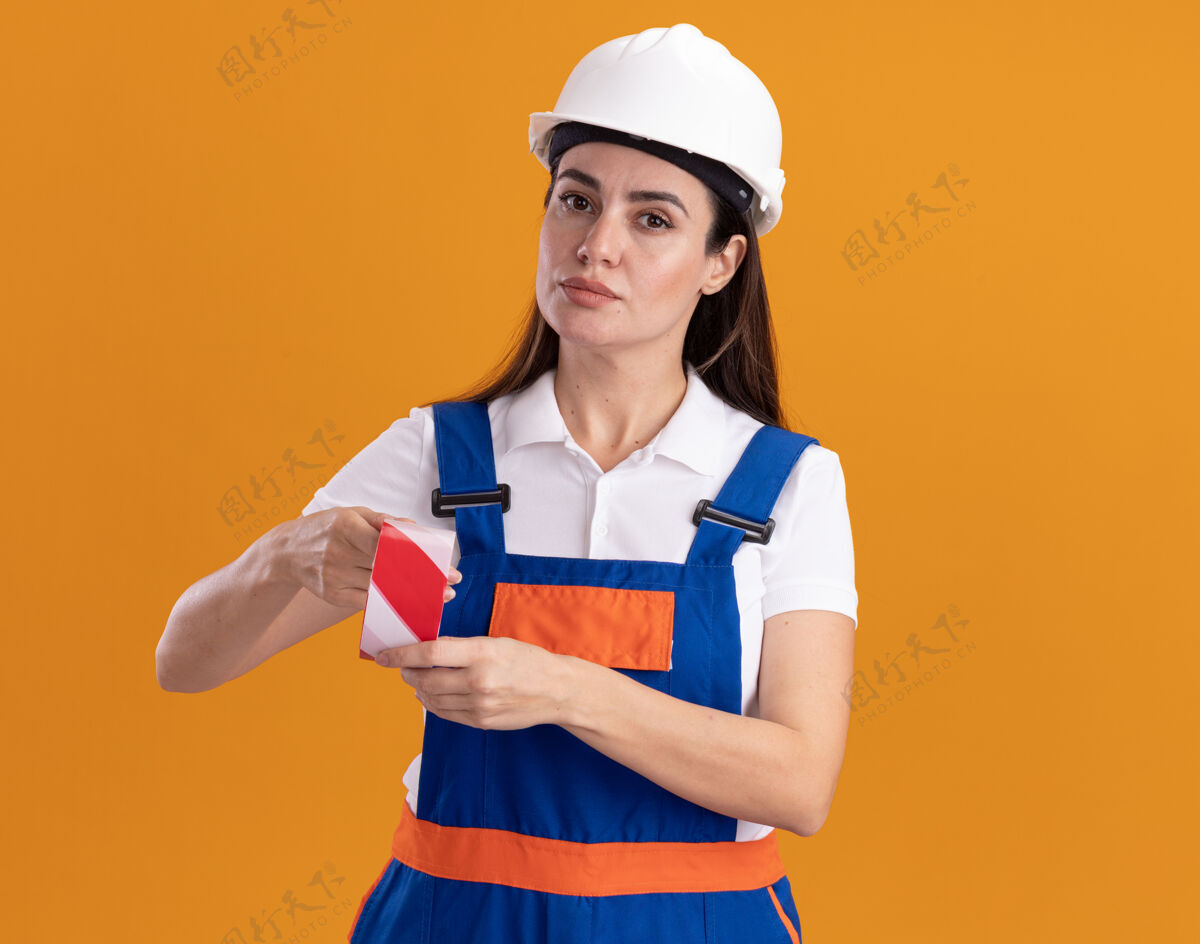 持有自信的年轻建筑妇女穿着制服拿着隔离在橙色墙上的管道胶带建设者磁带信心
