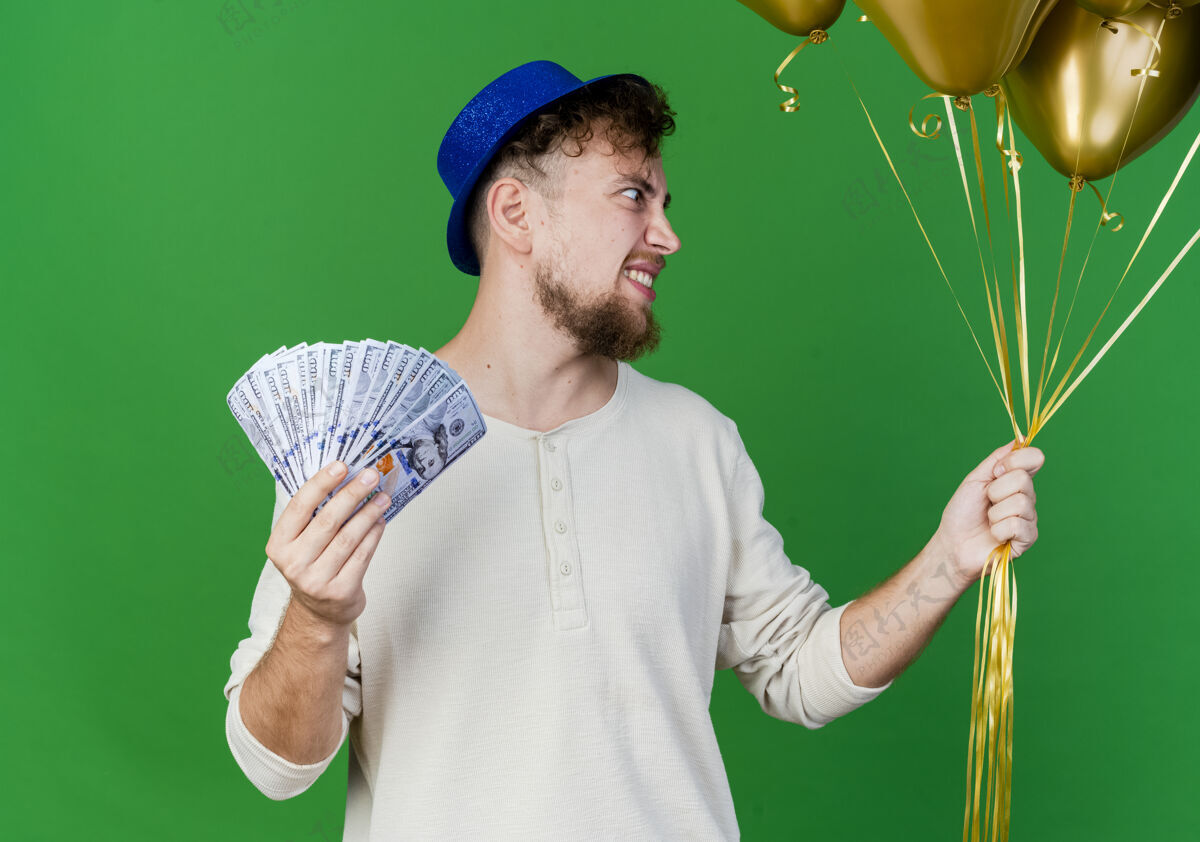 钱面带微笑的年轻帅哥斯拉夫党的家伙戴着党的帽子拿着气球和钱看一边孤立的绿色背景气球微笑绿色