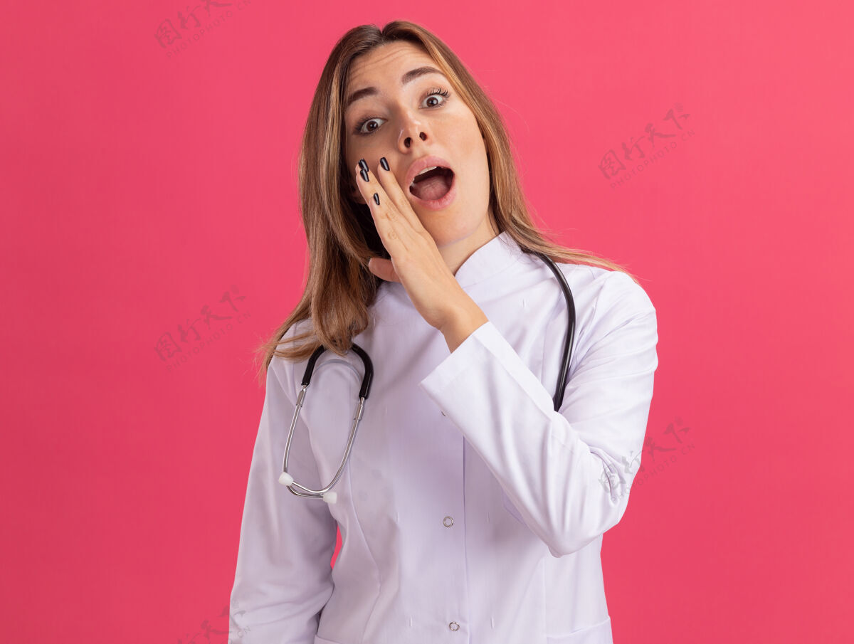耳语惊讶的年轻女医生穿着医用长袍 听诊器在粉红色的墙上低语年轻穿听诊器