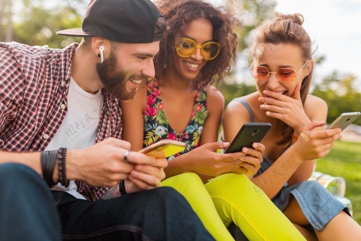 年轻快乐的年轻朋友们坐在公园里用智能手机微笑 男人和女人一起玩微笑朋友友谊