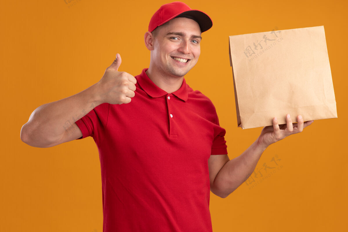 微笑笑容满面的年轻送货员穿着制服 戴着帽子 拿着纸食品包 竖起大拇指 孤立地站在橙色的墙上送货抱着纸张