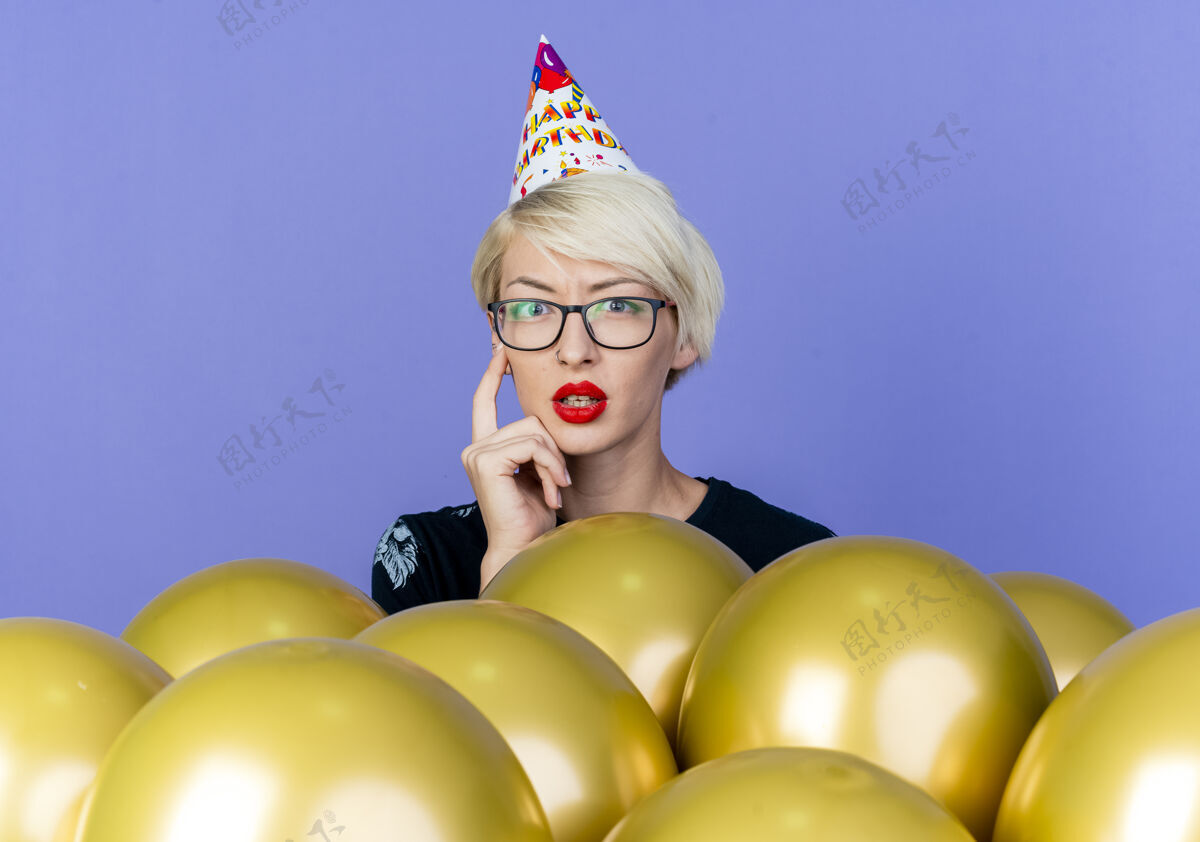 金发体贴的年轻金发派对女孩戴着眼镜和生日帽站在气球后面抚摸着脸看着紫色背景上孤立的相机派对生日紫色