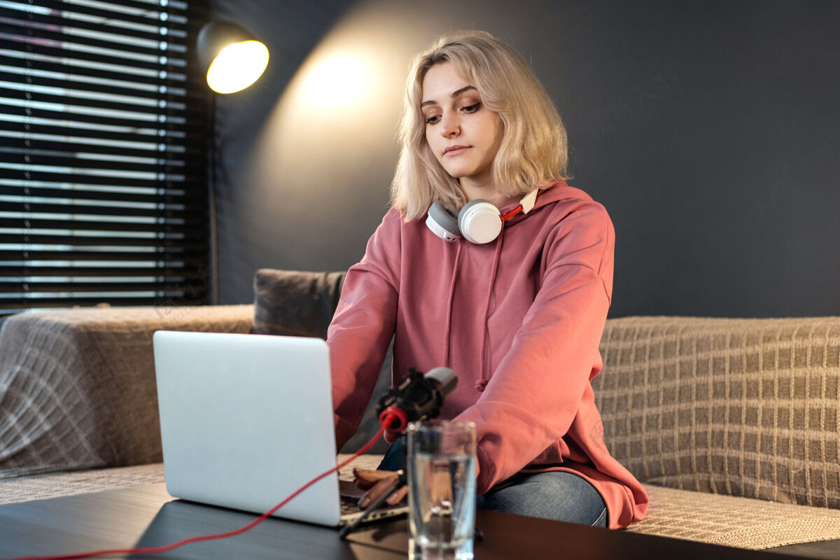 互联网年轻的内容创作者金发女孩戴着耳机在桌上的笔记本电脑上工作 带着相机工作女人播客