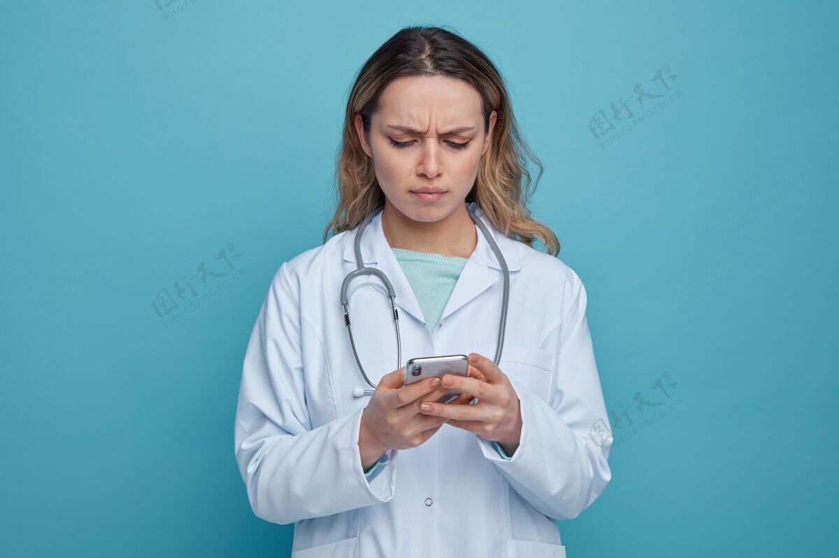 脖子皱眉的年轻女医生穿着医用长袍 用手机在脖子上绕着听诊器女性医生电话
