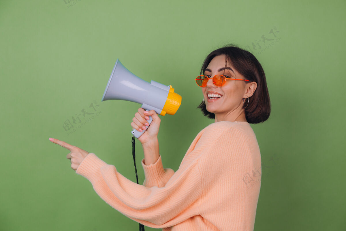 领导年轻女子戴着休闲的桃色和橙色眼镜毛衣 隔离在绿橄榄色的墙上 快乐地用扩音器尖叫 用食指指着左边的复制空间沟通新闻扩音器