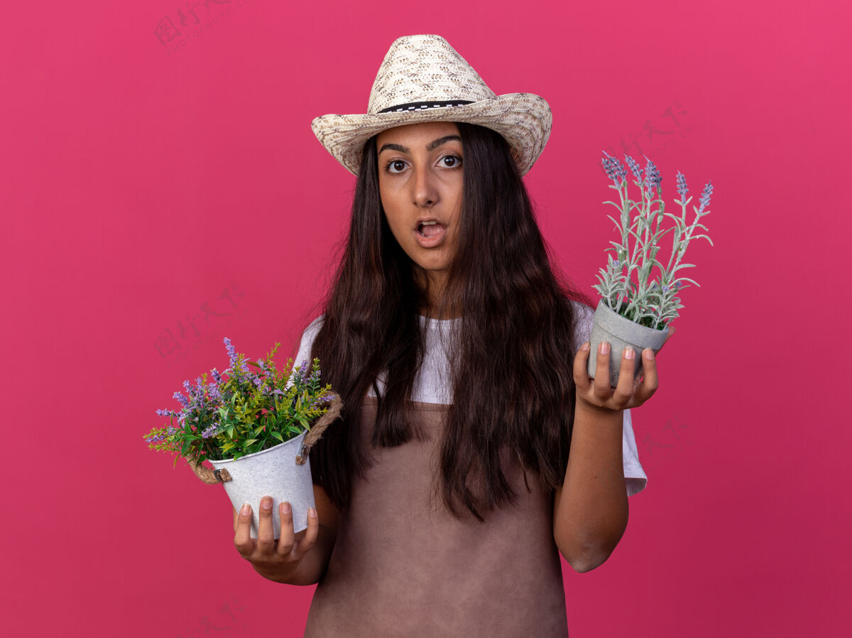 锅穿着围裙 戴着夏帽 手里拿着盆栽植物的年轻园丁女孩站在粉色的墙上 惊讶又惊奇围裙惊喜立场