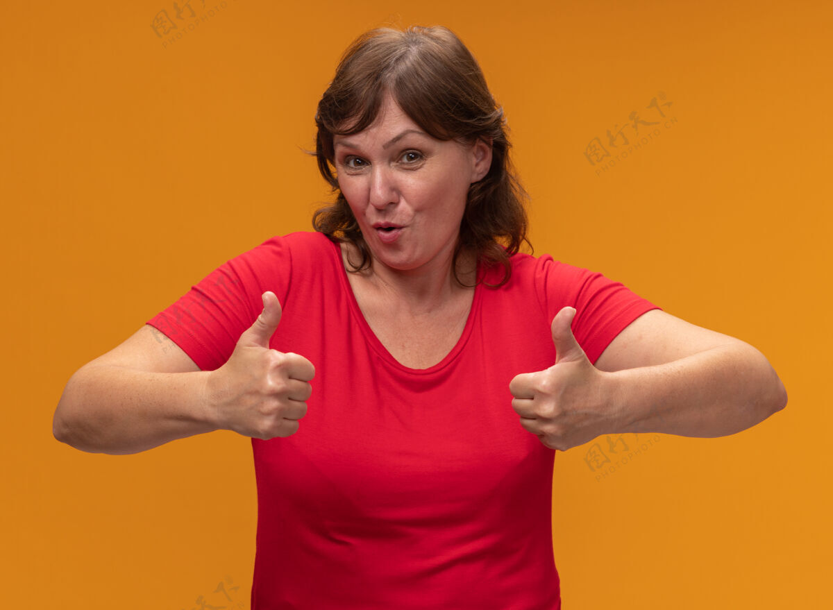 中间身着红色t恤的中年妇女站在橙色的墙上微笑着竖起大拇指拇指站立女人