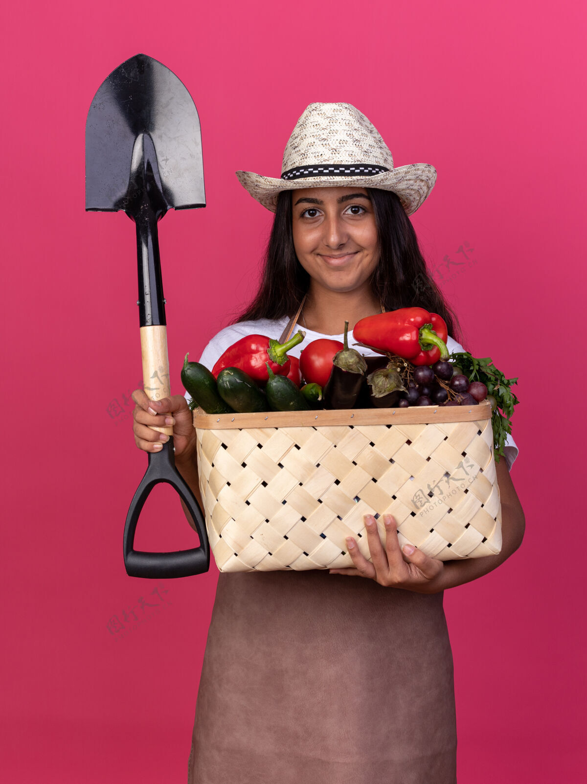 板条箱快乐的年轻园丁女孩 围着围裙 戴着夏帽 手里拿着装满蔬菜和铲子的箱子 脸上带着微笑站在粉红色的墙上铲子帽子花园