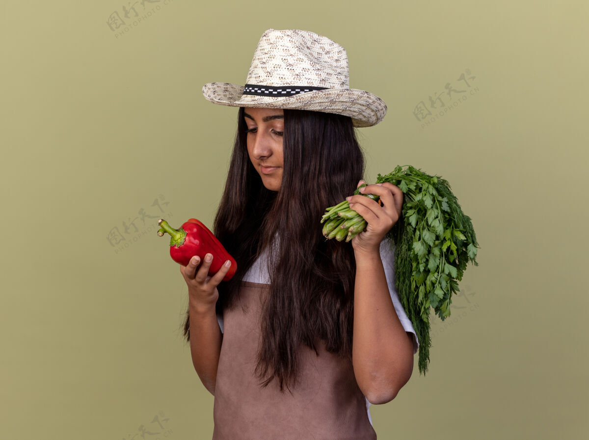 立场戴着围裙和夏帽的快乐的年轻园丁女孩 手里拿着新鲜的红甜椒和新鲜的草药 饶有兴致地看着站在绿墙上的胡椒兴趣围裙胡椒