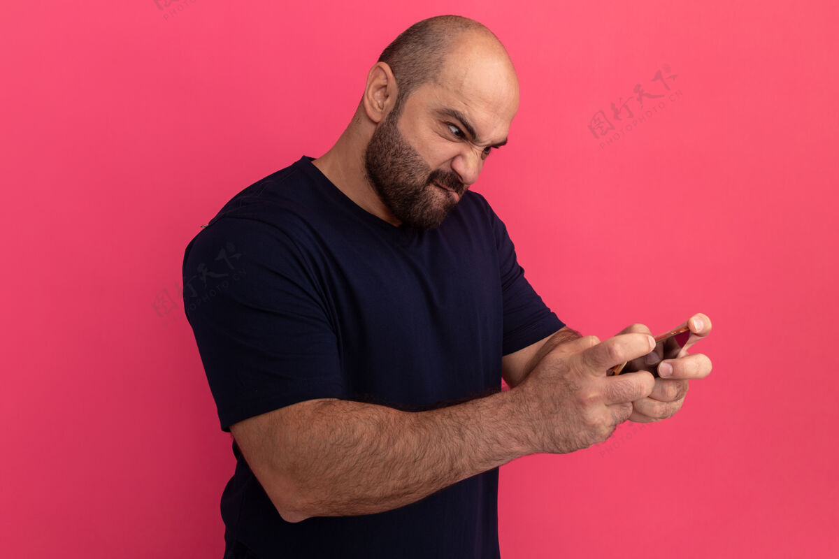 站着一个留着胡子的男人穿着海军蓝t恤 用智能手机玩游戏 站在粉色的墙上显得既恼火又兴奋兴奋游戏烦恼