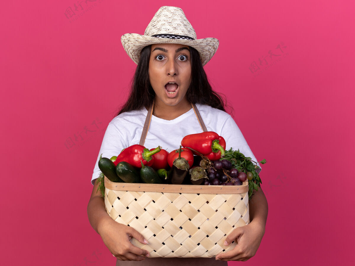 惊人年轻的园丁女孩围着围裙 戴着夏帽 手里拿着装满蔬菜的板条箱 站在粉色的墙上 既惊讶又惊奇帽子立场板条箱