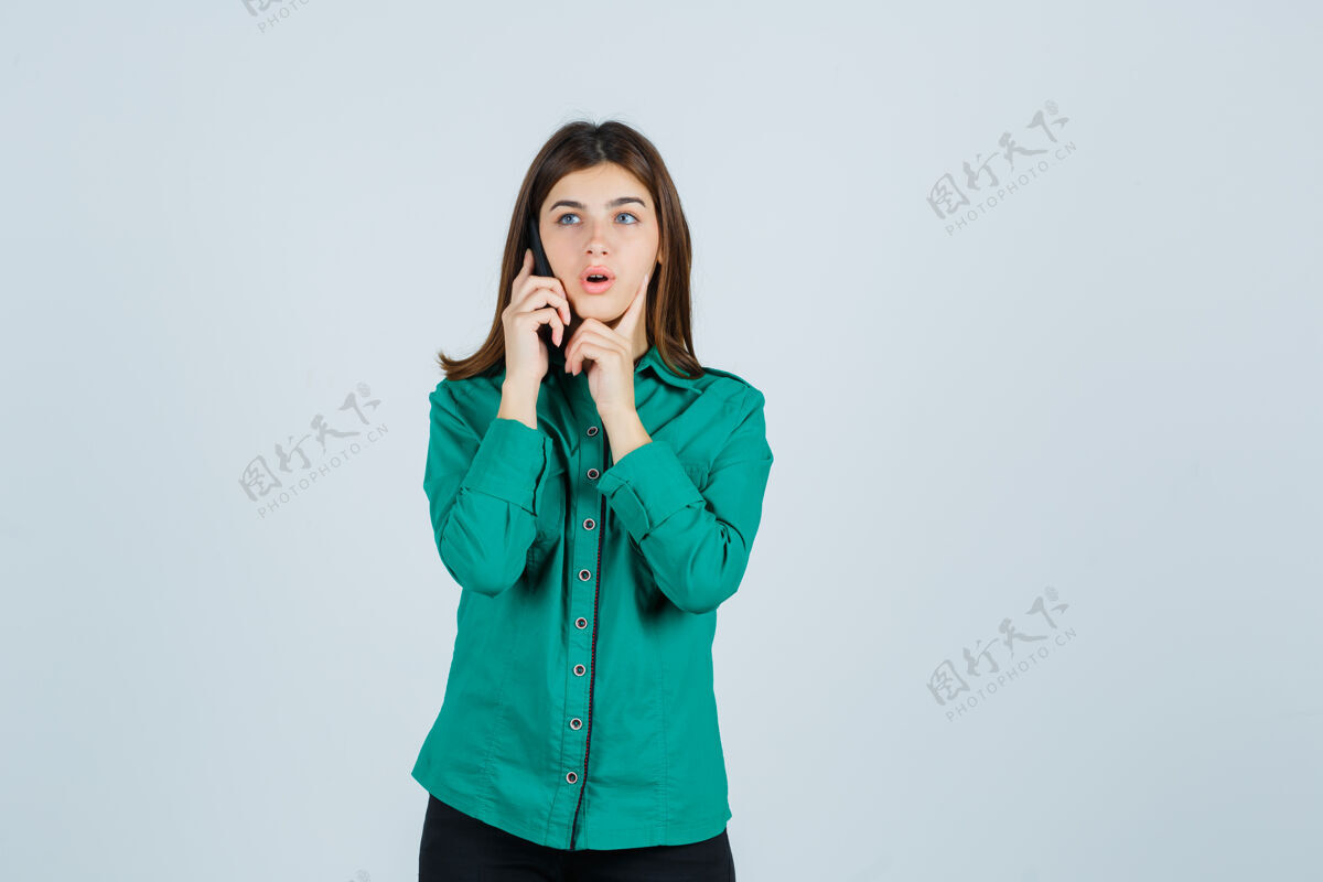 人年轻女孩在打电话 把食指放在下巴下面 穿着绿色上衣 黑色裤子 看起来很惊讶前视图女性面部手指
