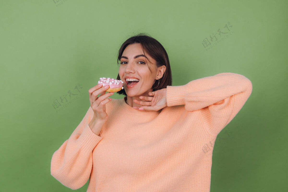 食物穿着休闲桃色毛衣的年轻时尚女性 隔离在绿色橄榄色墙壁上 粉色甜甜圈快乐复制空间女性肖像甜甜圈