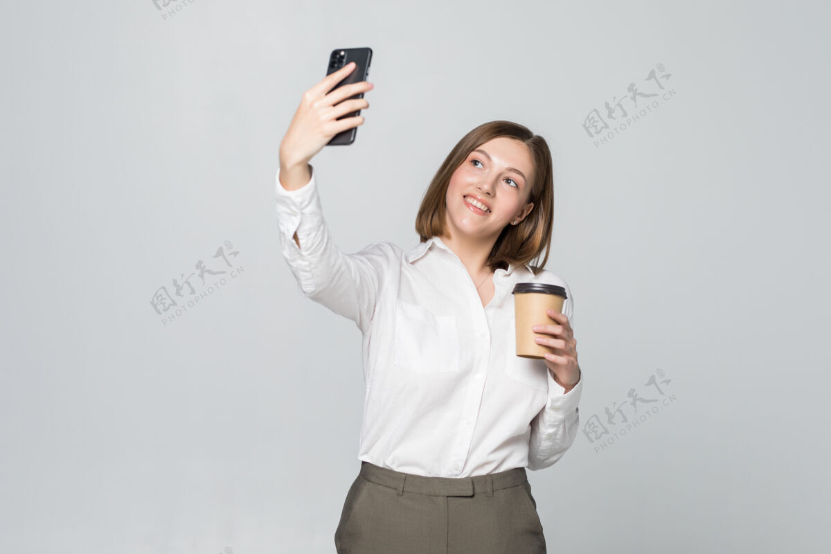 时尚身着正装的女商人手持外卖咖啡站在灰色墙壁上用手机自拍的照片年轻工作正式