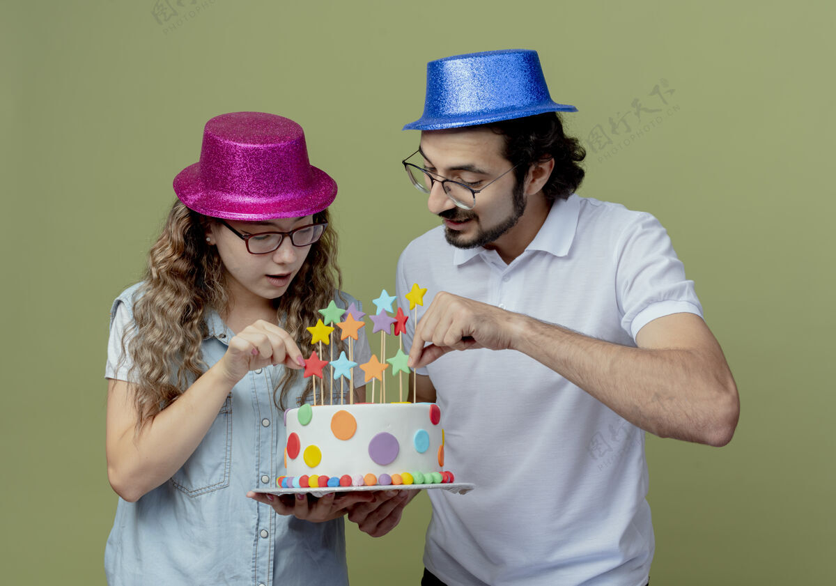 橄榄色高兴的年轻夫妇戴着粉红色和蓝色的帽子拿着生日蛋糕看着蛋糕情侣生日