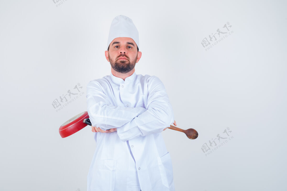 十字架年轻的男厨师手拿煎锅和木勺 双手交叉站在那里 身穿白色制服 看上去很自信 正对着前方制服厨师而