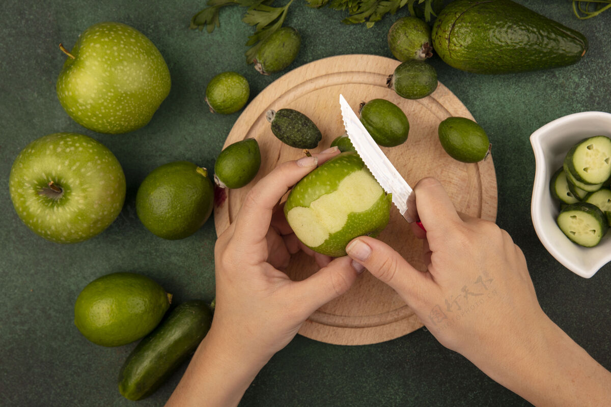 苹果俯视图：女性用刀在木制厨房板上剥一个新鲜的绿色苹果 在绿色的表面上隔离着酸橙 红豆和绿色的苹果农业板饮食