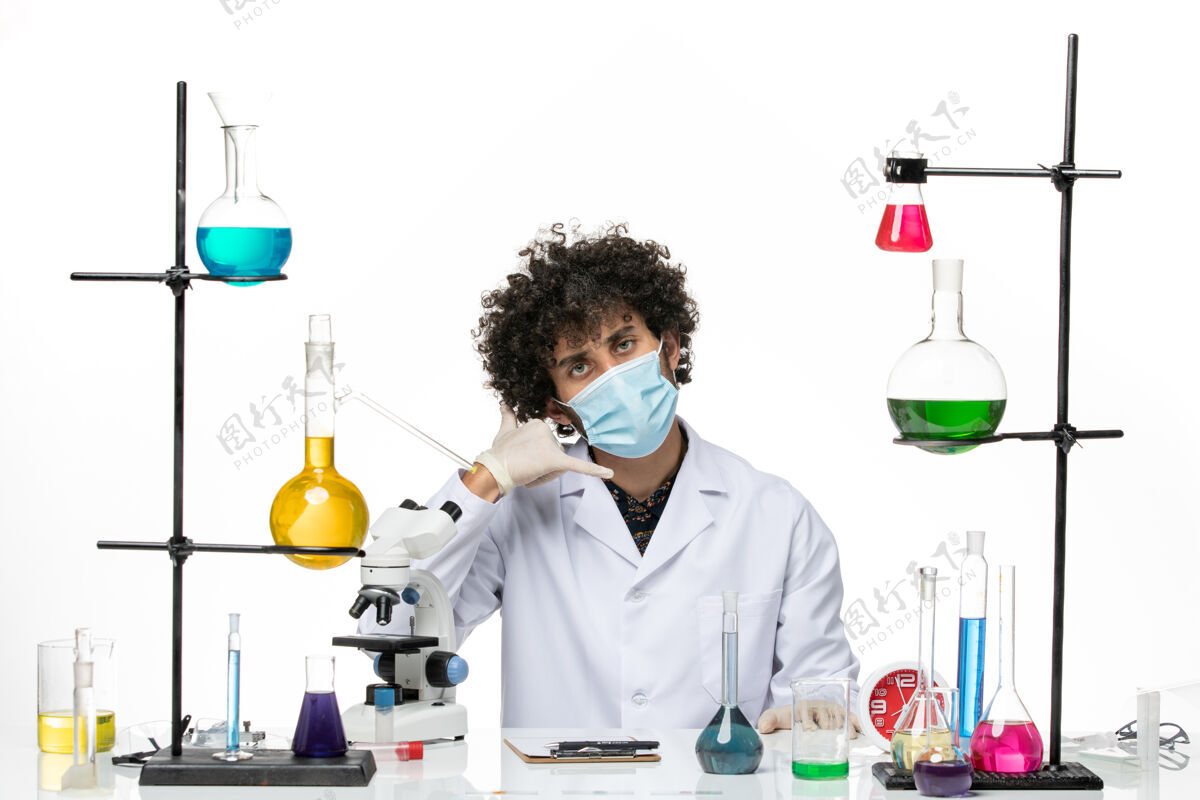 男性正面图：男药剂师穿着医疗服 戴着口罩 坐在不同的溶液中 在白色的空间里摆姿势解决方案化学男性化学家