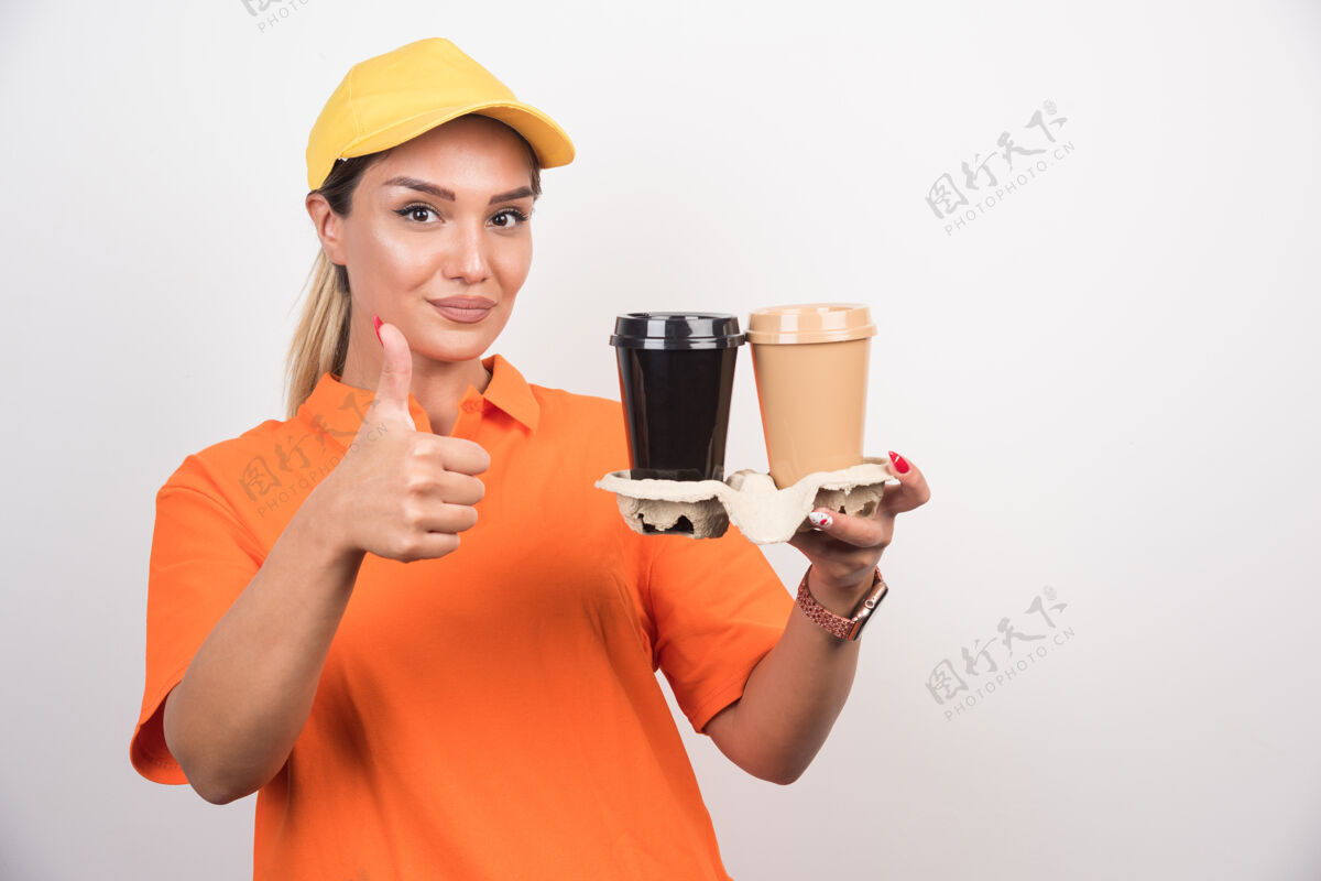外卖金发女信使拿着两杯咖啡 在白墙上竖起大拇指快递女人塑料杯