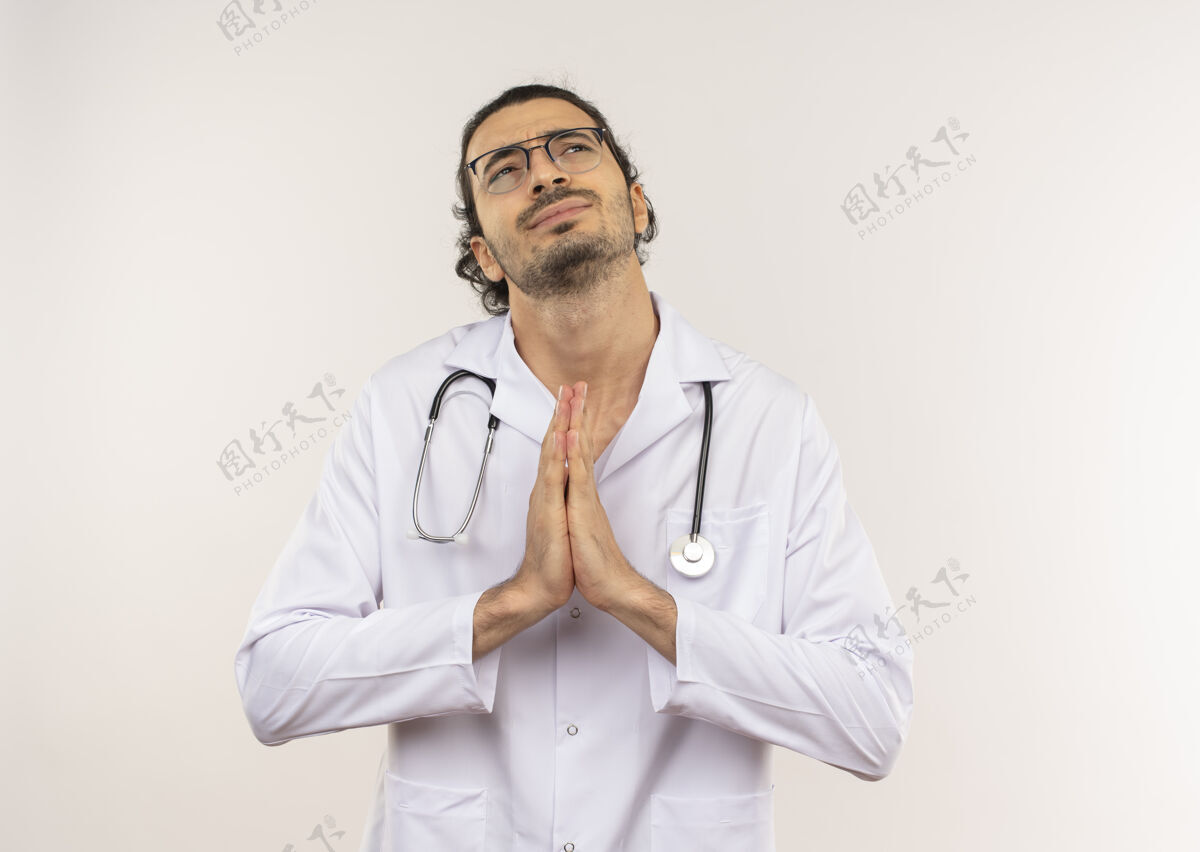 显示仰望着戴着眼镜 身穿白色长袍 手持听诊器的年轻男性医生 显示出祈祷的姿态眼镜关心长袍