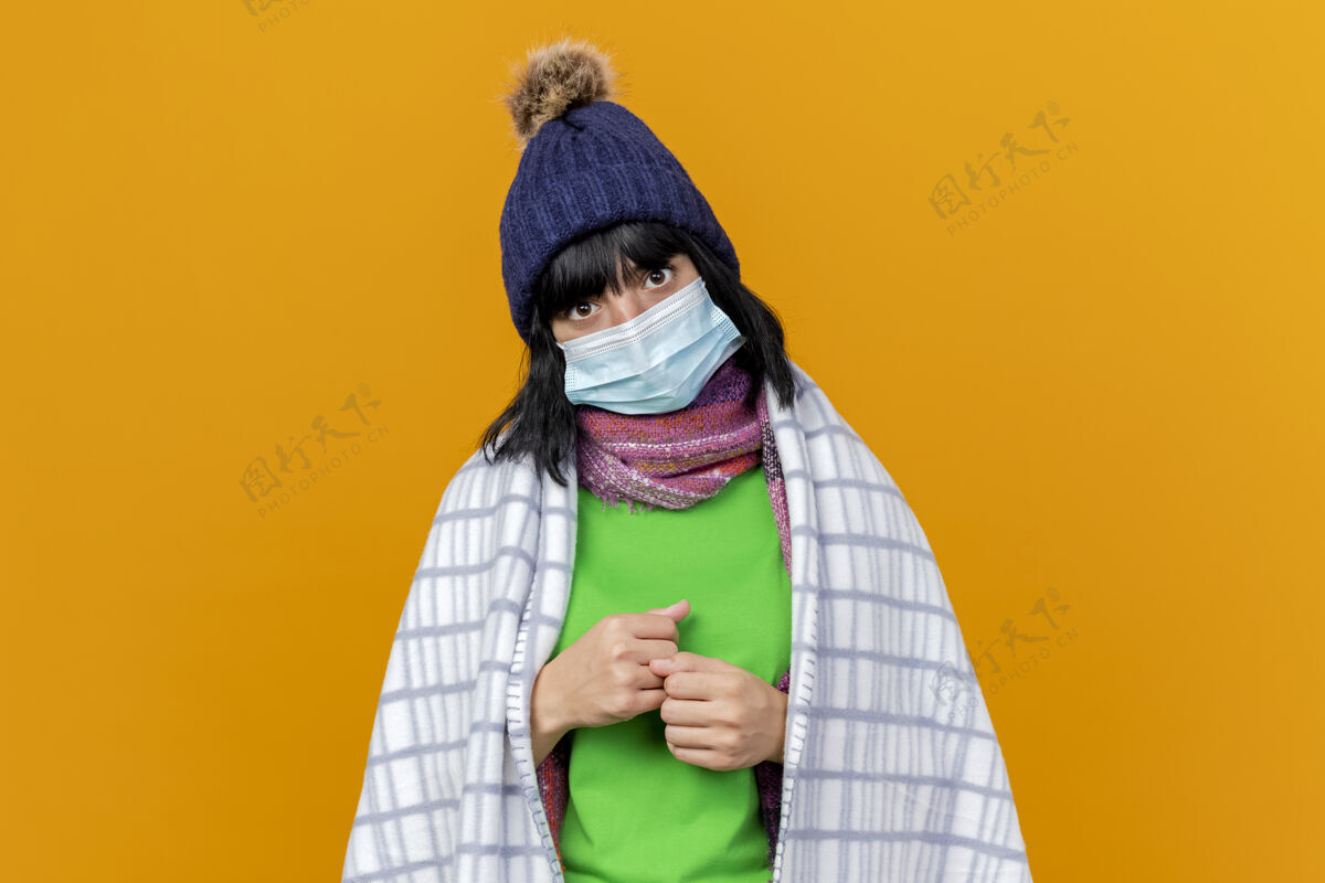 格子年轻生病的高加索女孩戴着冬天的帽子和围巾 戴着方格布的面具 紧握着拳头 隔离在橙色的墙上 留着复制空间穿年轻握紧