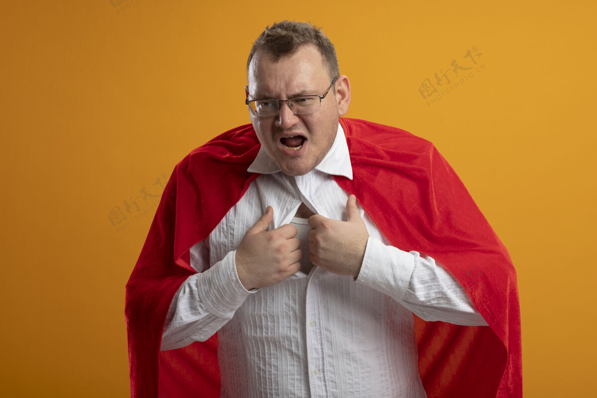 斗篷愤怒的成年斯拉夫超级英雄 穿着红色斗篷 戴着眼镜 抓着他的衬衫 试图脱下它 看着橙色墙上孤立的一面成人橙色男人