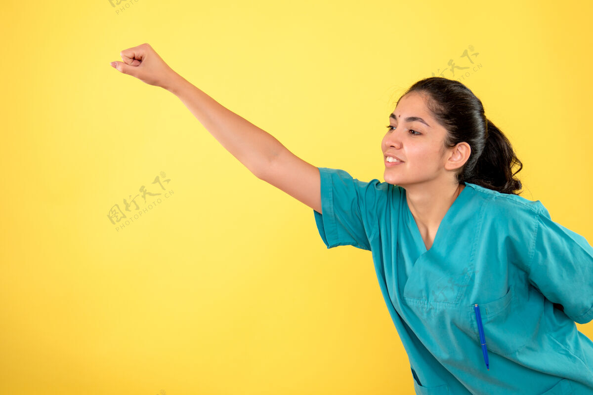 前面黄色墙壁上的超级英雄姿势的年轻女医生的正面图快乐模型漂亮
