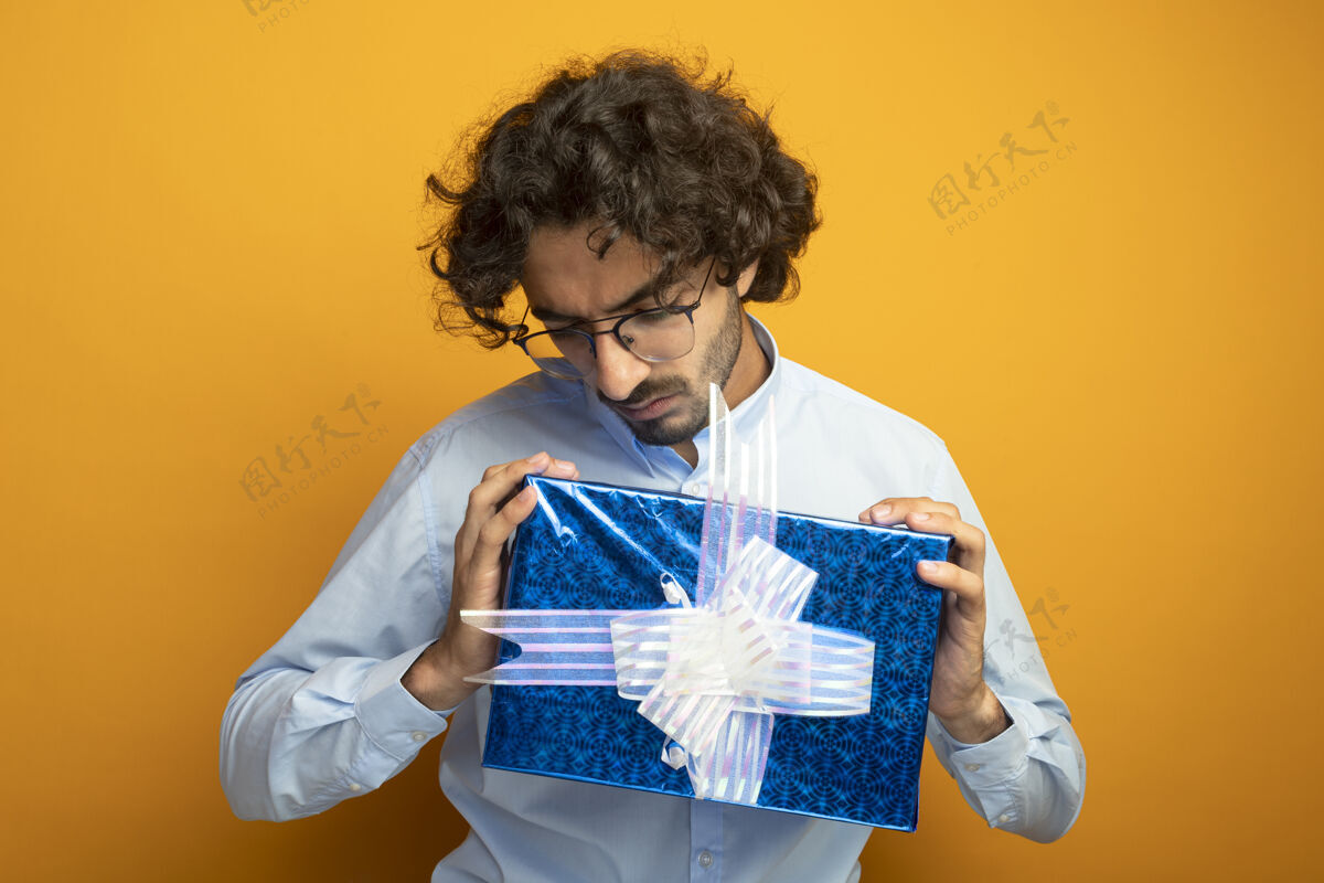 男人年轻英俊的白种人戴着眼镜拿着礼物包看着隔离在橙色墙上抱橙色包