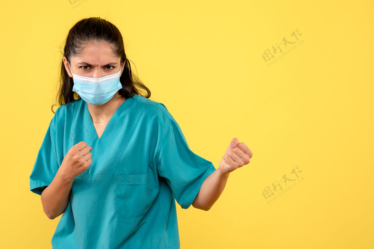 医疗戴着医用面罩的漂亮女医生的正面图 黄色墙壁上有拳头支架药健康