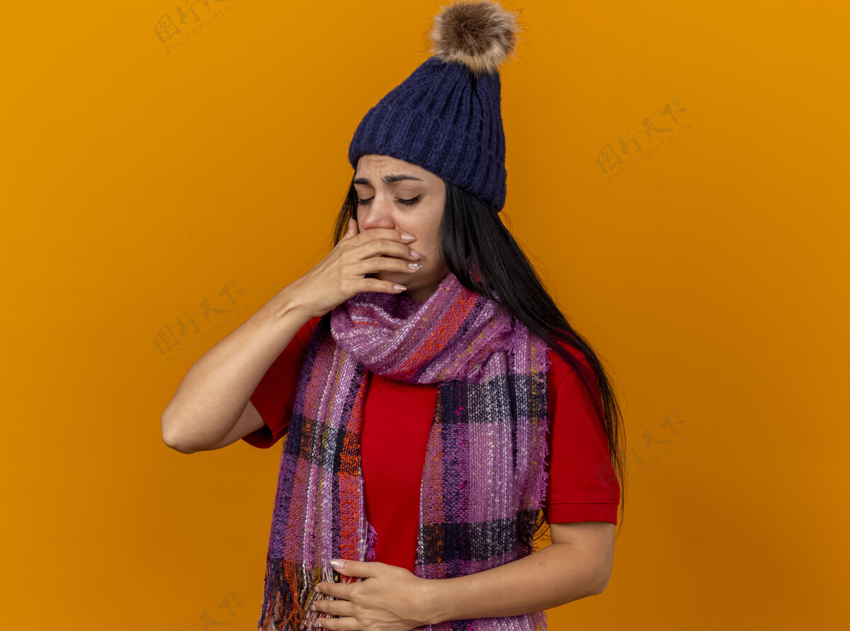 封闭疼痛的年轻白种人生病的女孩戴着冬天的帽子和围巾 手放在肚子上和嘴上 准备打喷嚏 闭上眼睛 隔离在橙色的墙上 留着复印空间穿腹部疼痛