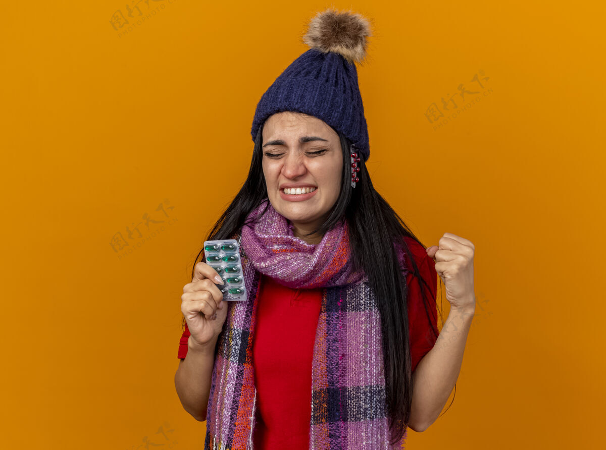 握紧恼怒的年轻白种人生病的女孩戴着冬天的帽子和围巾拿着一包胶囊和帽子下的其他包紧握着拳头闭着眼睛隔离在橙色的墙上复制空间拳头胶囊橙色