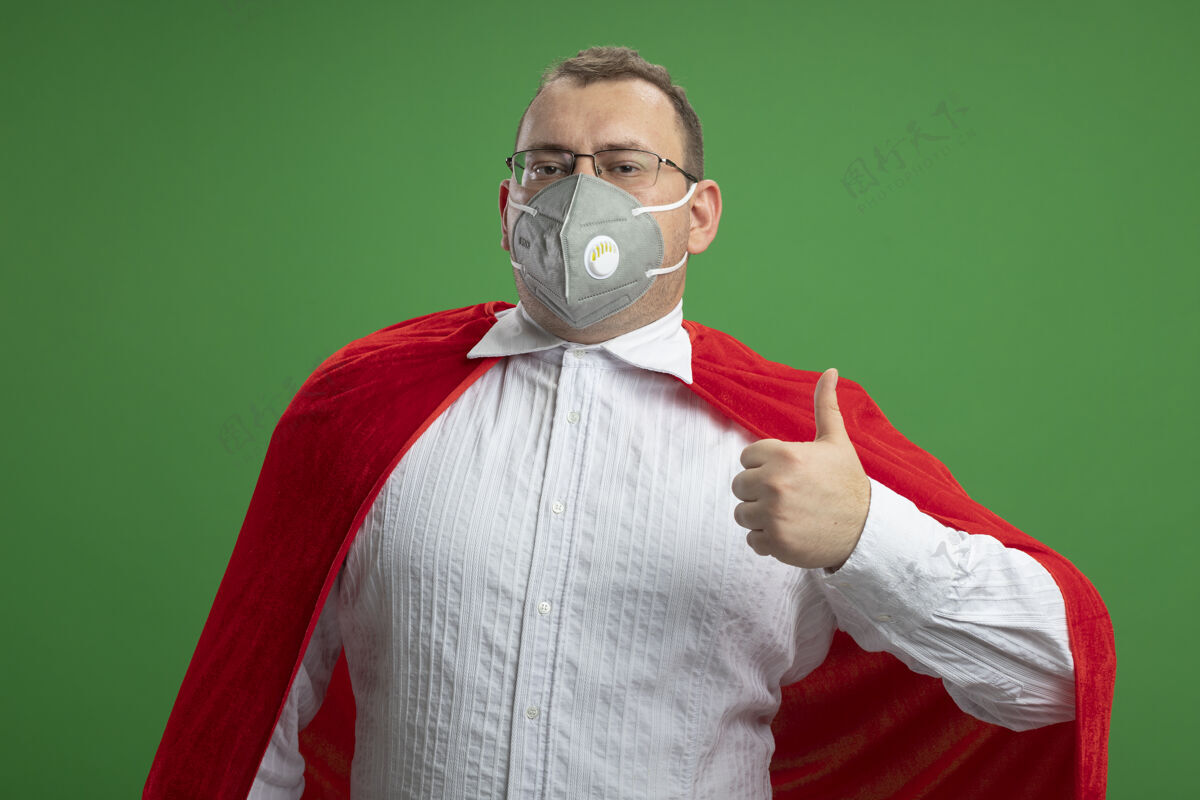 超级英雄自信的成年斯拉夫超级英雄身穿红色斗篷 戴着眼镜和防护面具 在绿色的墙上孤立地竖起大拇指面具拇指穿着