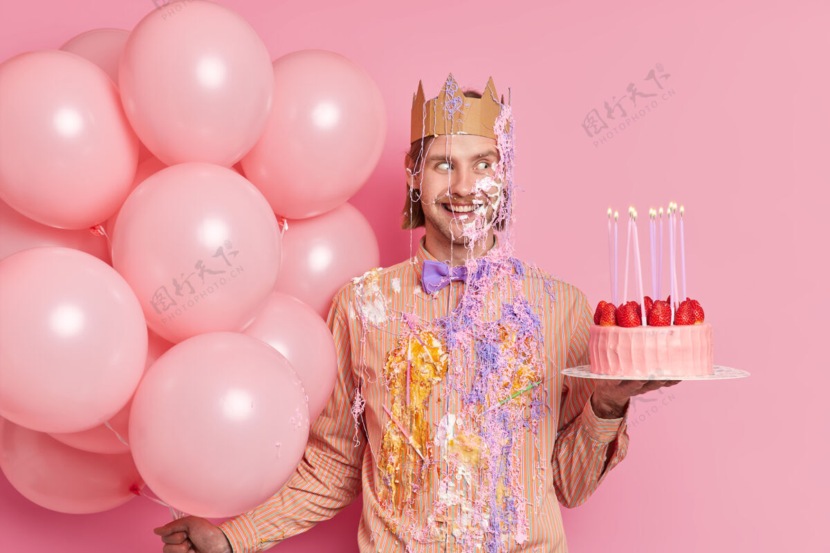 男性室内拍摄的英俊开朗的男子庆祝周年纪念涂抹奶油举行美味蛋糕和气球在生日派对上的乐趣隔离粉红色的墙壁请帅哥站