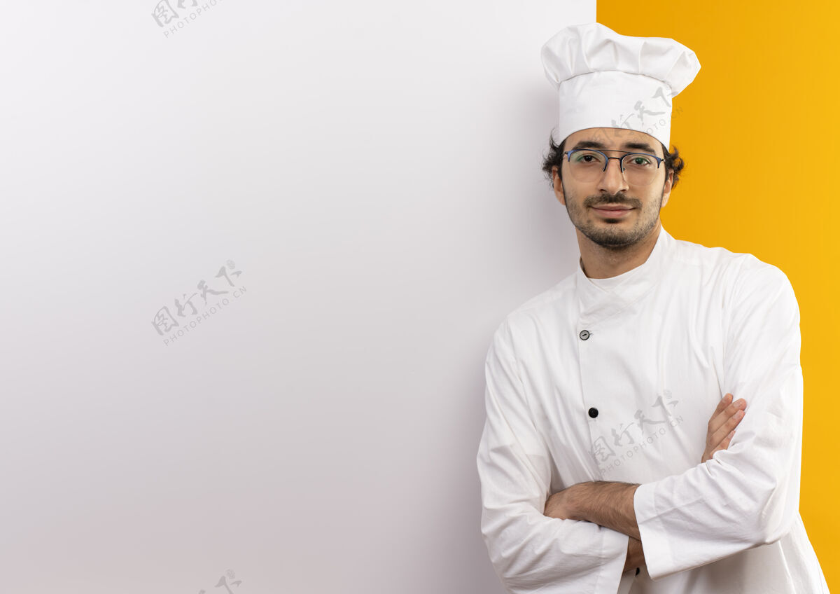 站立高兴的年轻男厨师穿着厨师制服和眼镜交叉双手站在白色的墙壁隔离在黄色的墙壁与复制空间男性交叉烹饪