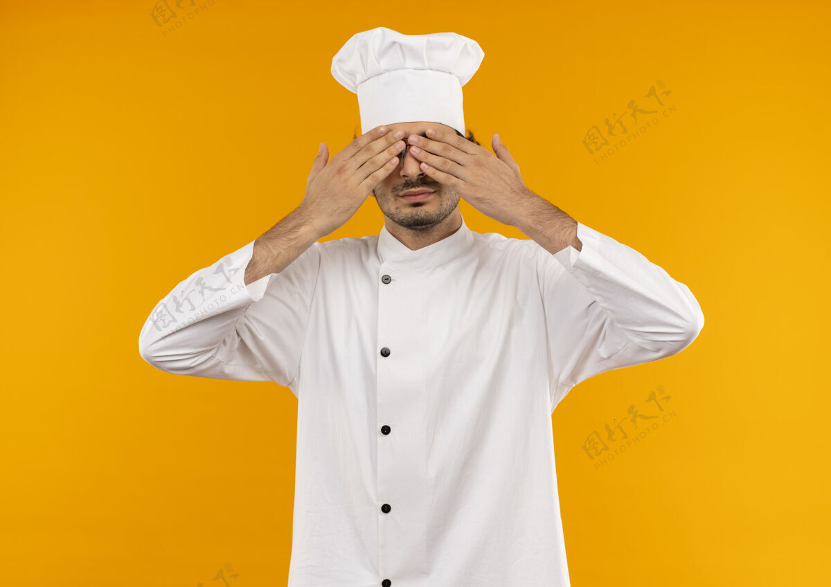 请穿年轻的男厨师穿着厨师制服 戴着眼镜 双手孤立在黄色的墙上 眼睛被遮住了制服年轻眼镜