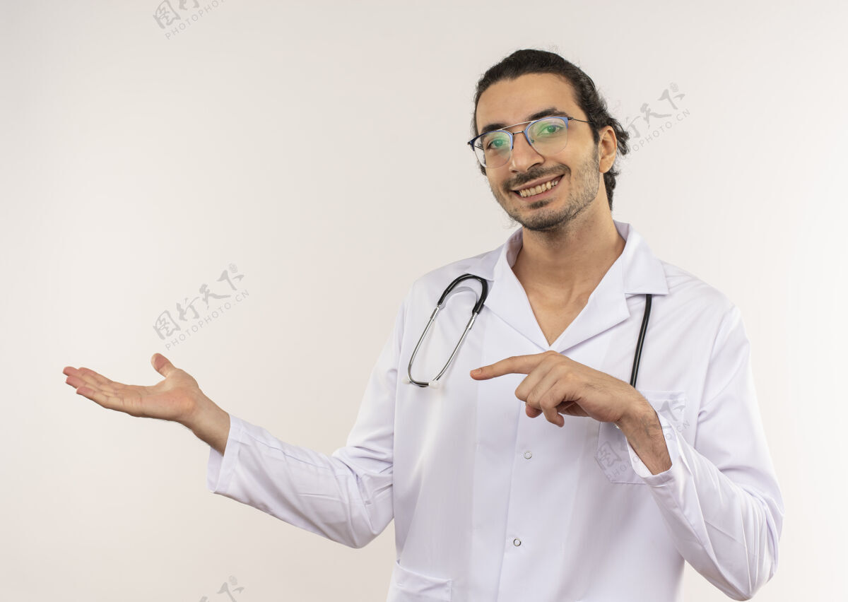 男性面带微笑的年轻男医生戴着眼镜 穿着白色长袍 手持听诊器 指着隔离的白墙上有复印空间的一侧眼镜长袍微笑
