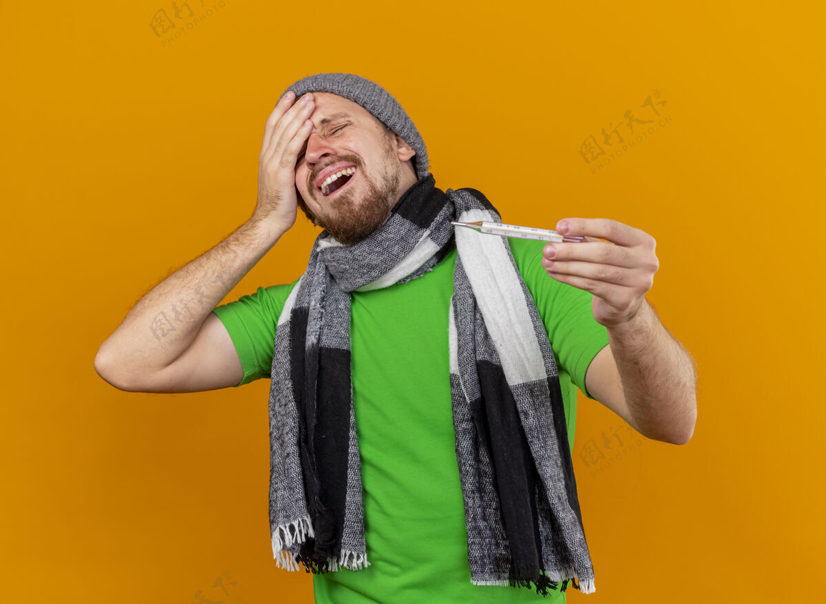 男人年轻英俊的斯拉夫病夫戴着冬天的帽子 戴着围巾 抱着温度计 闭着眼睛 摸着头 隔离在橙色的墙上疼痛橙色头