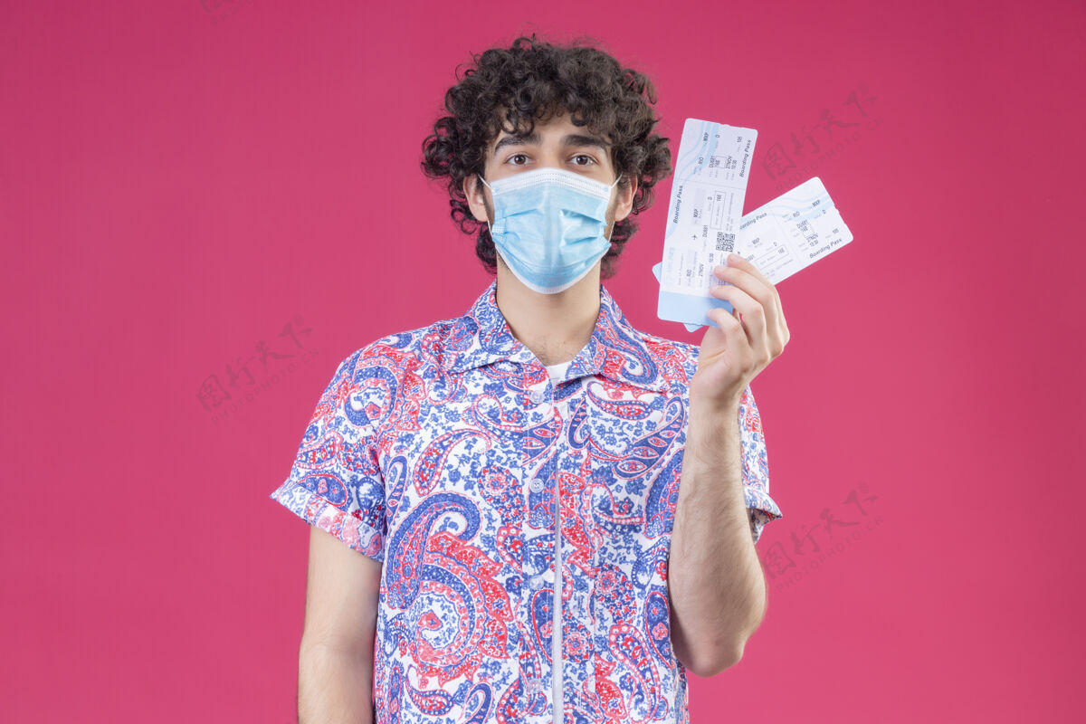 持有年轻帅气的卷发旅行者戴着医用口罩拿着飞机票站在隔离的粉色墙上 还有复印空间男人医疗面具