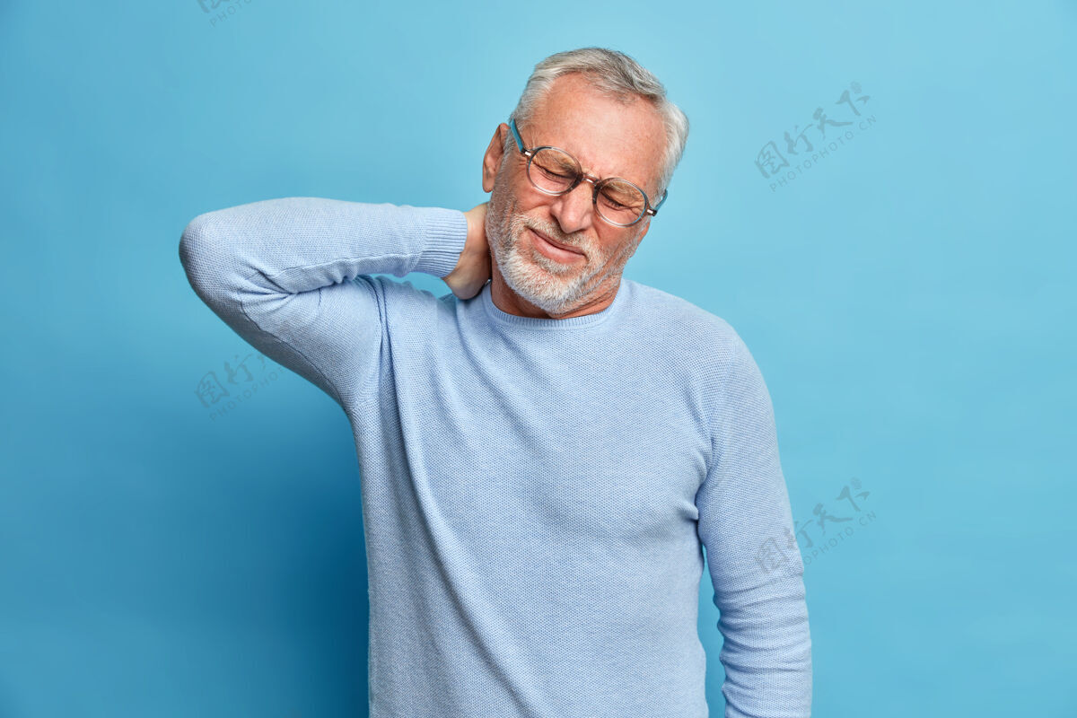 退休年迈疲惫的留着胡须的欧洲男人摸着脖子忍受着脖子上的疼痛歪着头做鬼脸痛苦的感觉需要按摩穿着长袖套头衫隔着蓝色的墙壁人疼痛触摸
