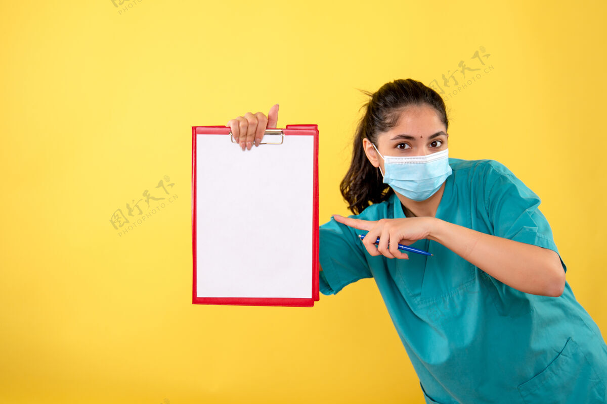 年轻女性年轻女性的前视图 医学面具指向黄色墙上的剪贴板人快乐微笑