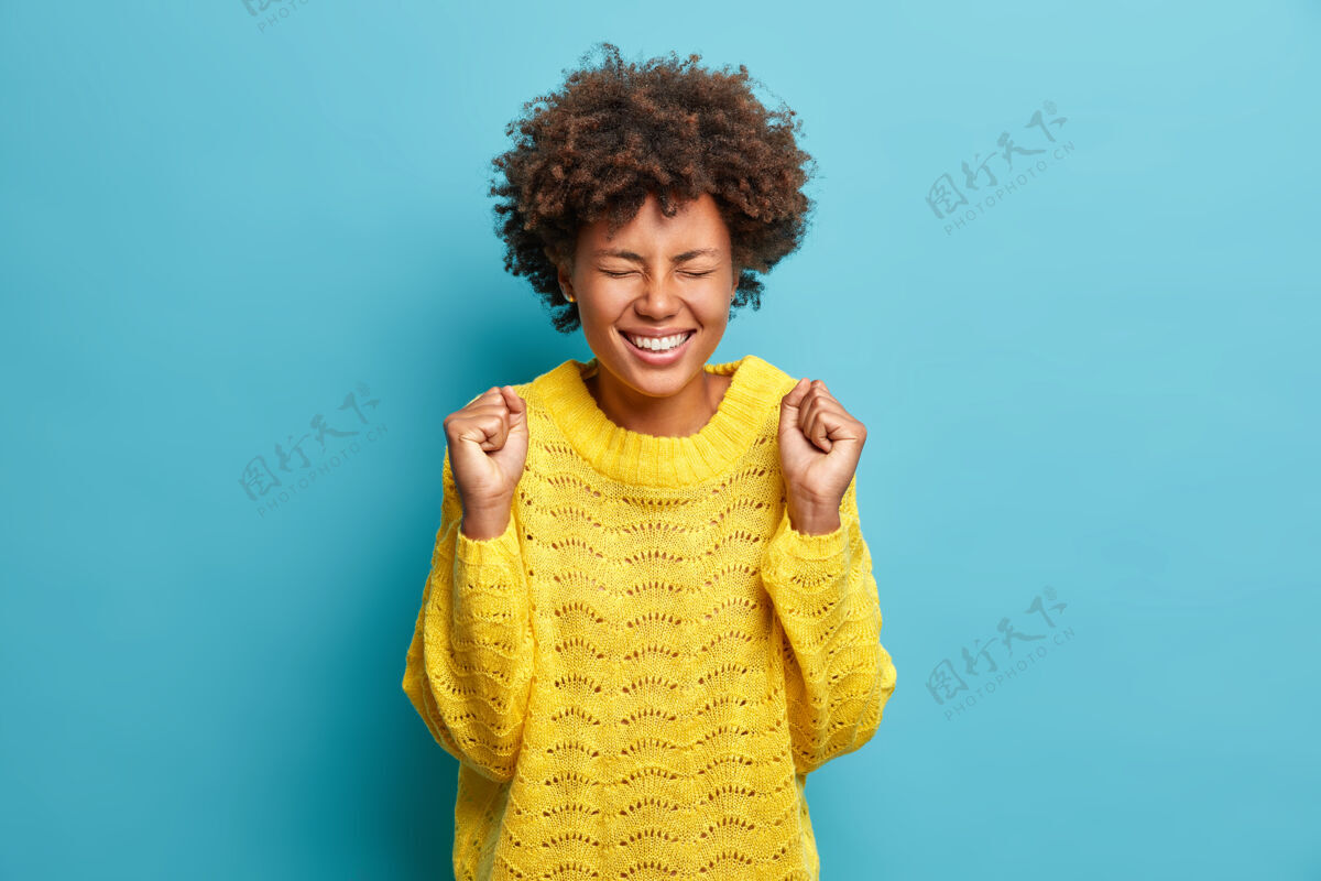 模特喜气洋洋的女人在收到好消息后欢呼并握紧拳头看起来喜气洋洋地穿上了针织的黄色毛衣有趣美丽头发