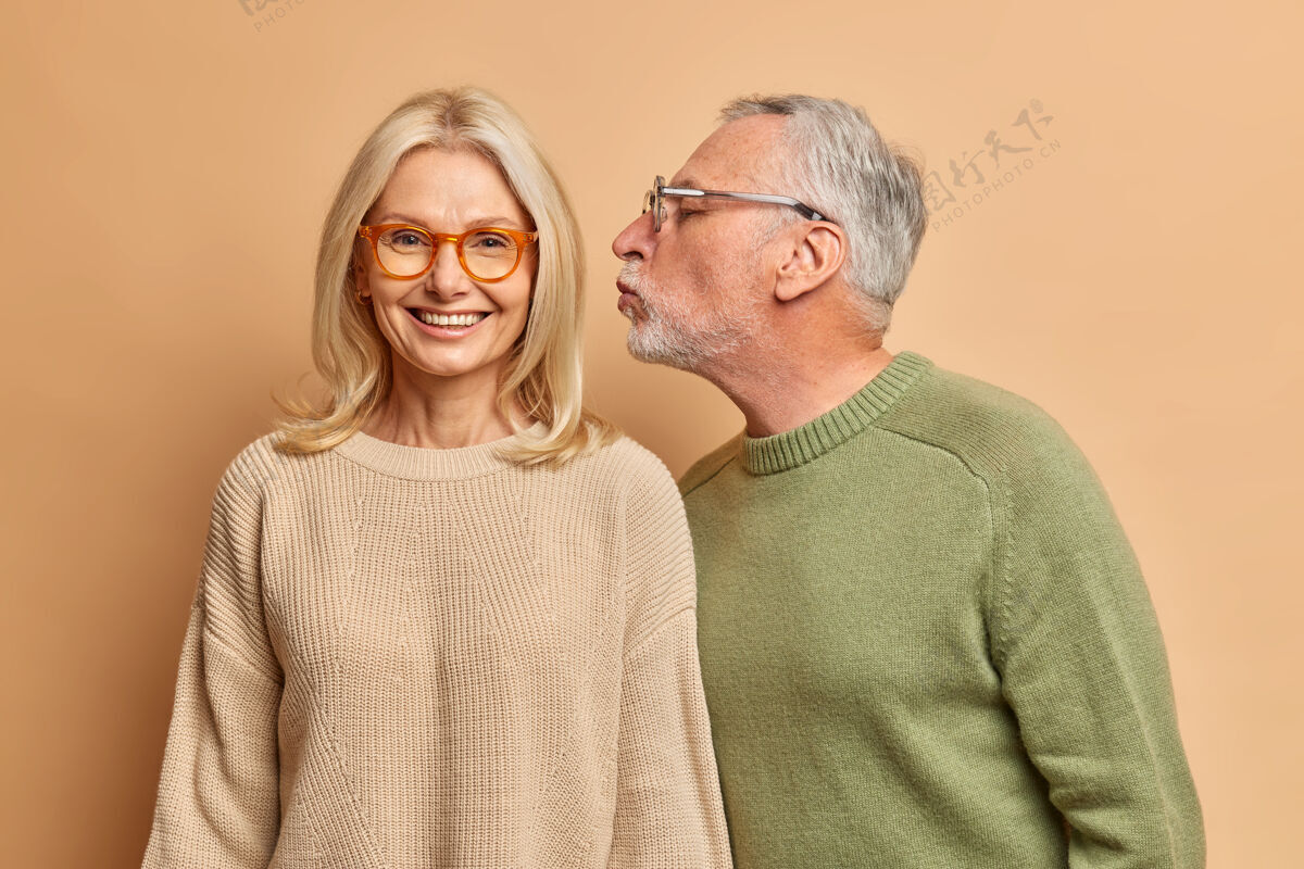 男性开朗的欧洲中年妇女微笑着 温柔地接受丈夫的亲吻 有着良好的关系 彼此相爱很久 隔着棕色的墙浪漫退休高兴