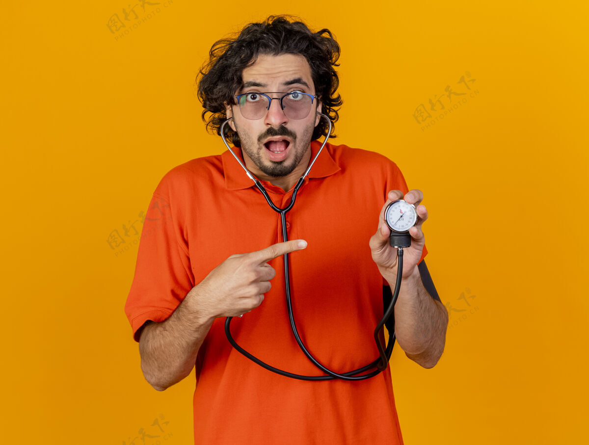 血压计印象深刻的年轻白种人病人戴眼镜和听诊器举行和指着血压计隔离在橙色墙上的复制空间疾病橙色男人