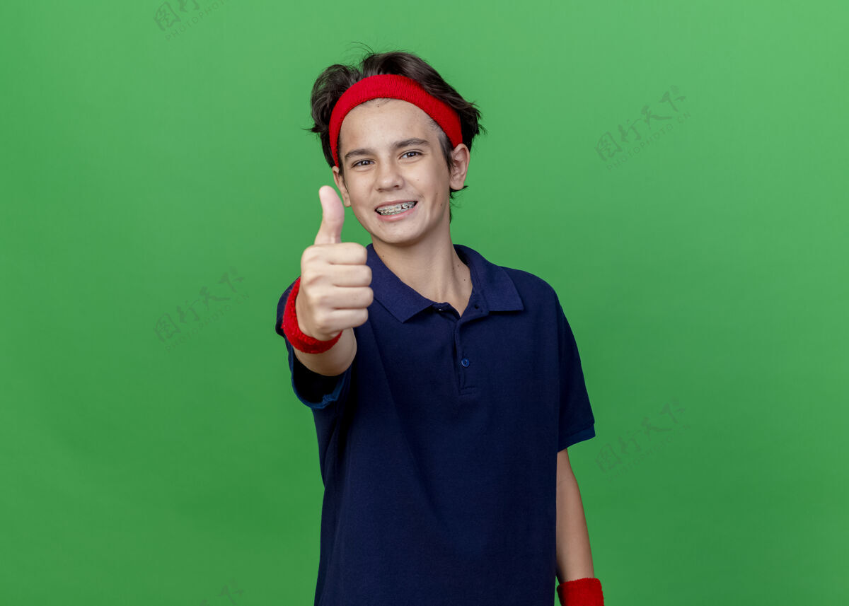帅气面带微笑的年轻帅气的运动男孩戴着头带和护腕 戴着牙套 大拇指向上竖立 隔离在绿色的墙上 留着复制空间拇指头带腕带
