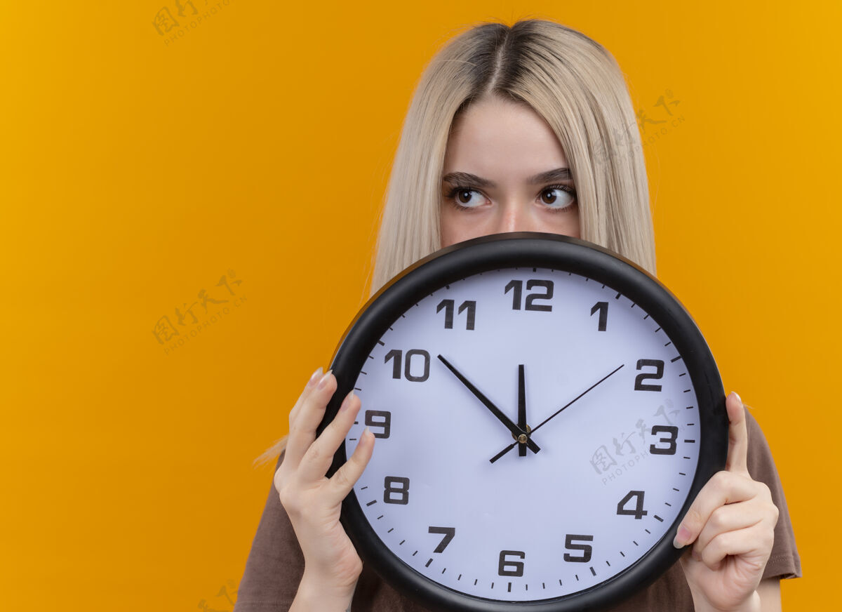 金发年轻的金发女孩拿着时钟躲在它后面 看着左边的橙色墙壁上的复制空间左边侧线女孩