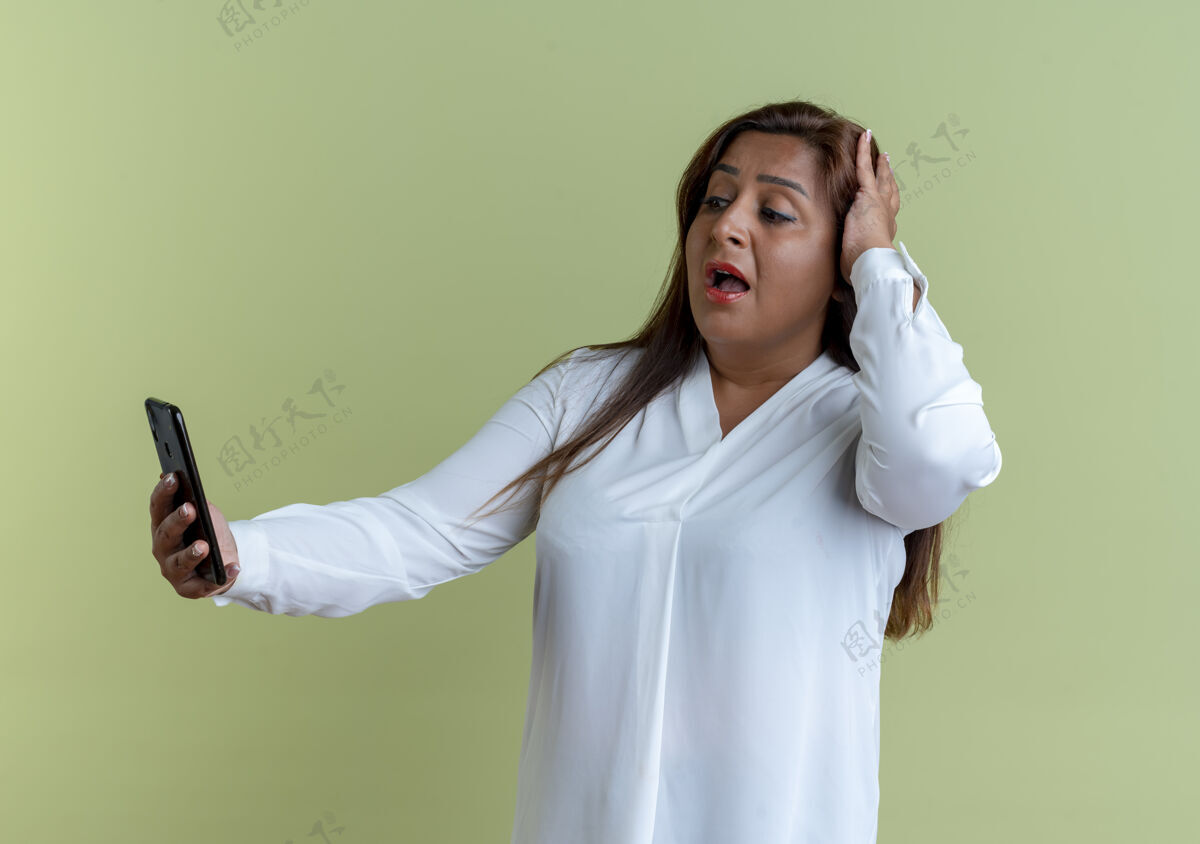 抓住惊讶的是 一个不经意的白人中年妇女拿着电话 看着电话 抓住了孤立在橄榄绿墙上的头头休闲橄榄