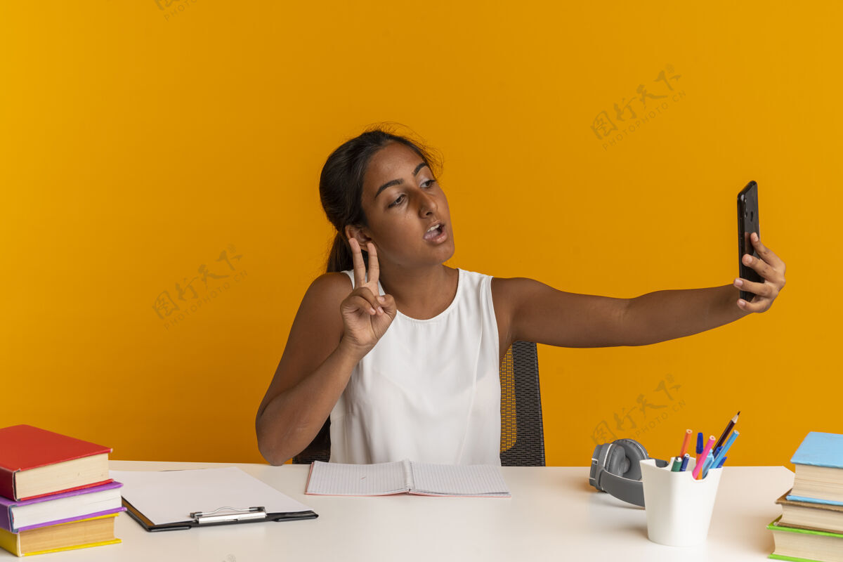 采取坐在书桌旁 拿着学习用具的小女孩高兴地自拍 在橘色的墙上表现出一种与世隔绝的和平姿态女学生工具书桌