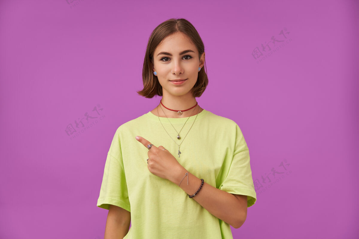 T恤女学生 一位棕发短发的年轻女士 用手指指着紫色墙上的复印处展示一个标志穿着绿色t恤 手镯和戒指女士20多岁黑发