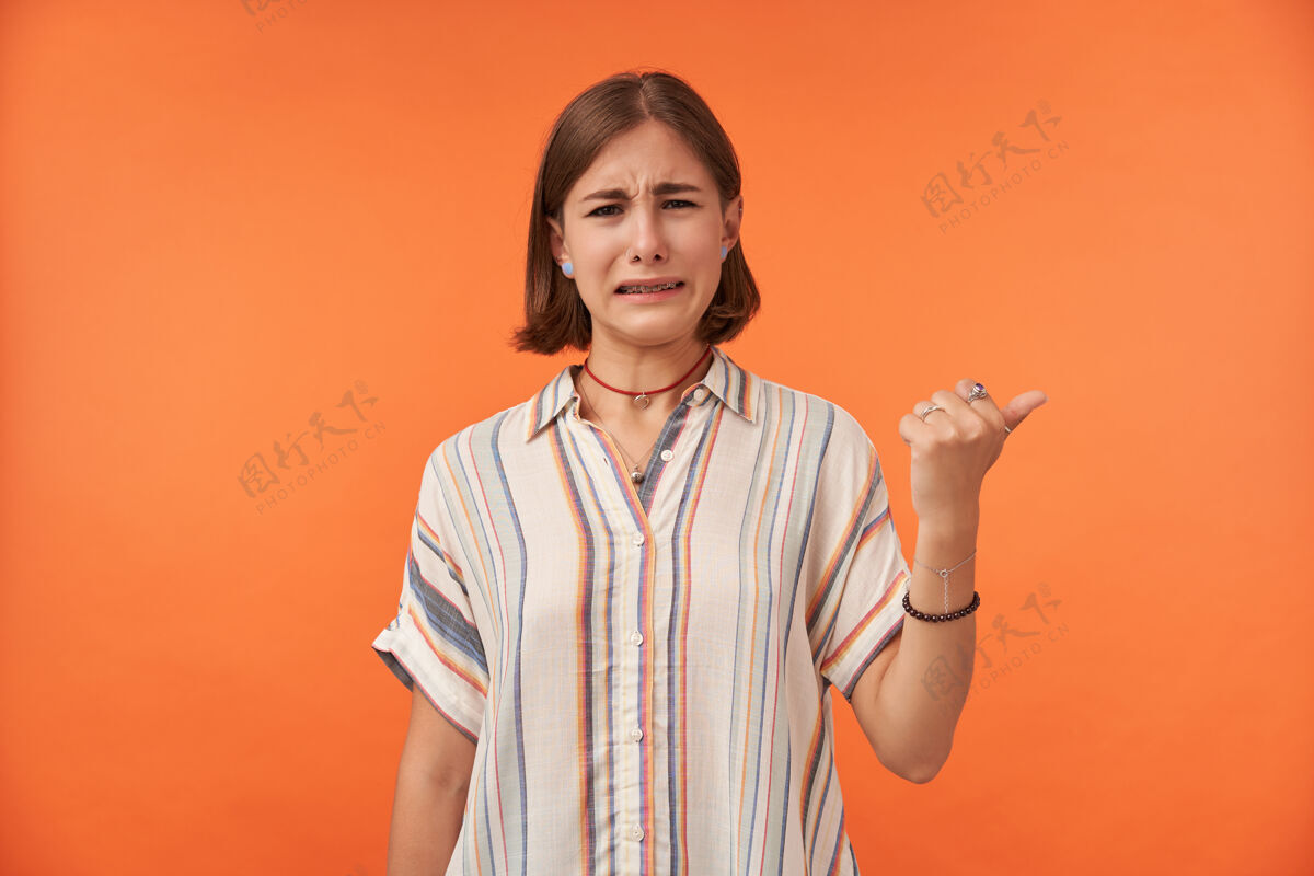 女人照片上是一个心烦意乱的女孩 她对自己看到的东西感到恶心 指着橘色墙上的复制空间的右边穿着条纹衬衫 项链 手镯和戒指年轻孤独不快乐