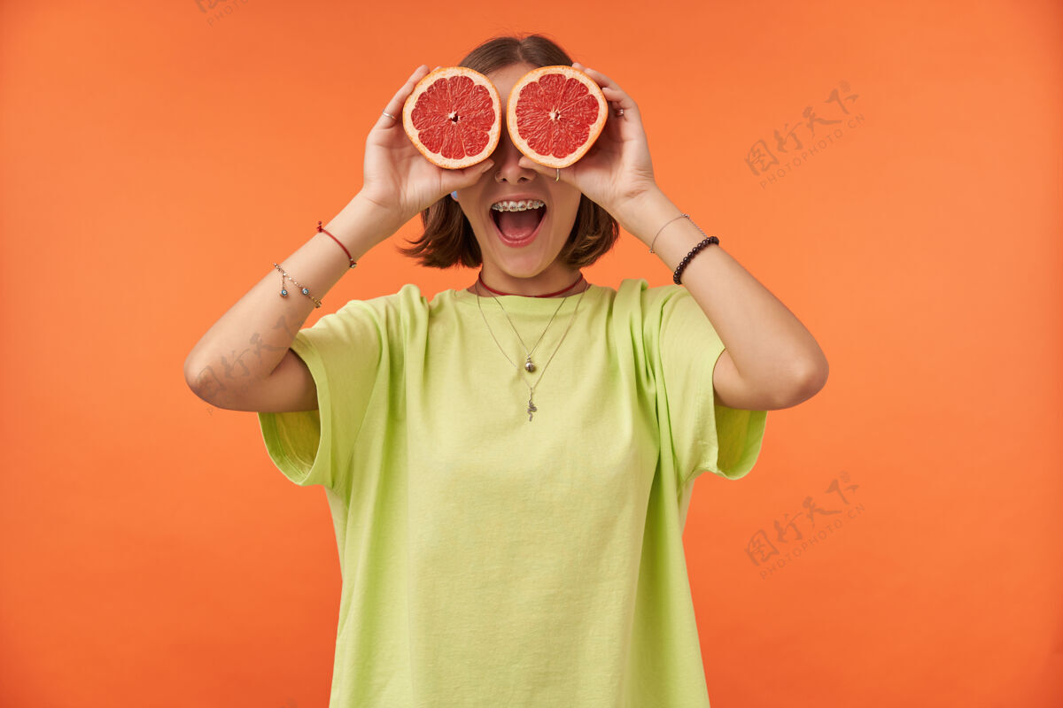 时髦女学生 一位年轻的女士 深色短发 眼睛上蒙着葡萄柚神情惊讶站在橙色的墙上穿着绿色t恤 戴着牙套和手镯20多岁背带热带